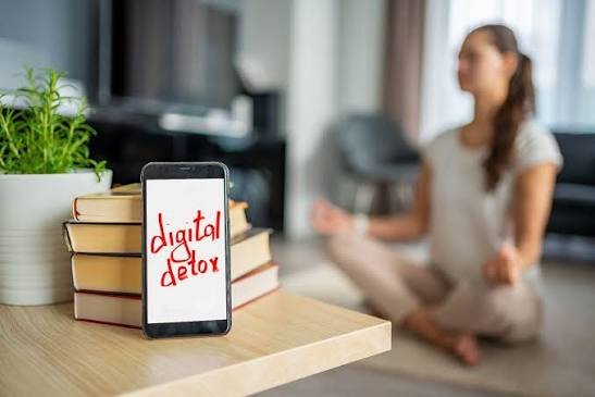 Smartphone with "digital detox" text on a stack of books, while a woman meditates in the background
