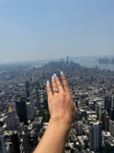 Angela Nikolau's hand showing an engagement ring extended over the New York City skyline seen from the top of the Empire State Building