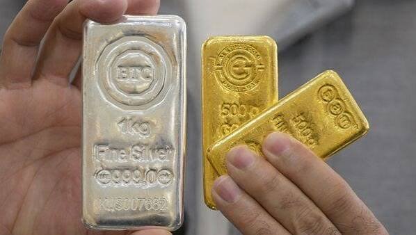 A hand holding a one kilogram fine silver bar next to two gold bullion bars representing today gold and silver investment rates
